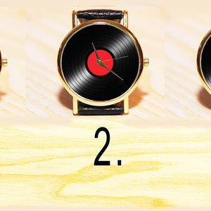 May include: Three gold-tone wristwatches with black leather bands and black vinyl record faces. The center of each watch face features a different colored circle: blue, red, and yellow.