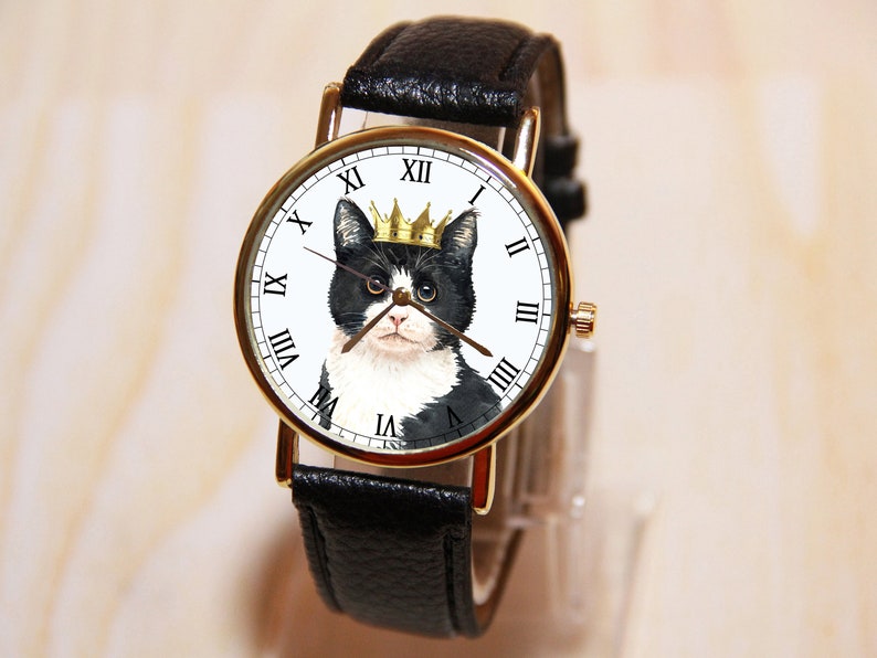 Op de afbeelding: Een polshorloge met een zwarte leren band en een gouden wijzerplaat waarop een zwart-witte kat met een gouden kroon is afgebeeld.