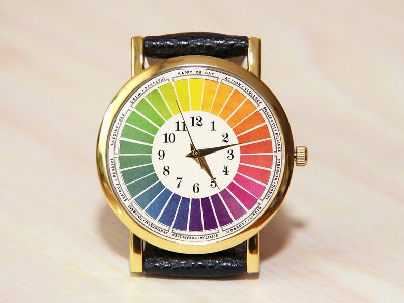 Puede incluir: Un reloj de pulsera dorado con una correa de cuero negro. La esfera del reloj tiene un fondo blanco con un dise&ntilde;o circular de colores y agujas de hora y minuto negras. Los n&uacute;meros del 1 al 12 est&aacute;n impresos alrededor del borde de la esfera del reloj.