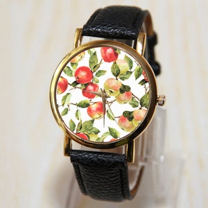 May include: A gold-tone wristwatch with a black leather band and a white face featuring a repeating pattern of red and yellow apples.