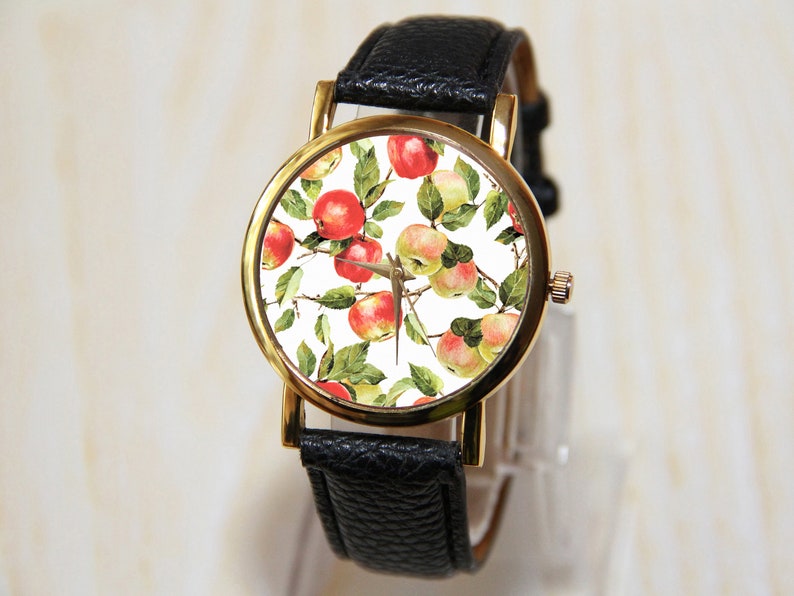 May include: A gold-tone wristwatch with a black leather band and a white face featuring a repeating pattern of red and yellow apples.