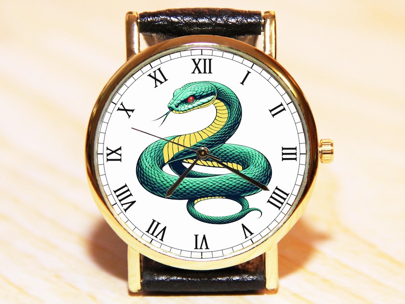 Puede incluir: Un reloj de pulsera dorado con una esfera blanca y una correa de cuero negra. La esfera presenta marcadores de hora en n&uacute;meros romanos y un dise&ntilde;o de serpiente verde.