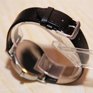 Puede incluir: Un reloj de pulsera plateado con una correa de cuero negro y una hebilla plateada. El reloj est&aacute; en un expositor transparente.