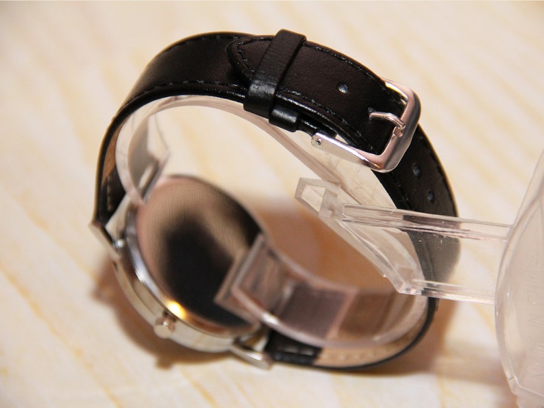 Puede incluir: Un reloj de pulsera plateado con una correa de cuero negro y una hebilla plateada. El reloj est&aacute; en un expositor transparente.