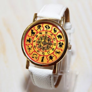 May include: A white wristwatch with a gold-tone case and a yellow face. The face features a zodiac design with black silhouettes of the astrological signs.