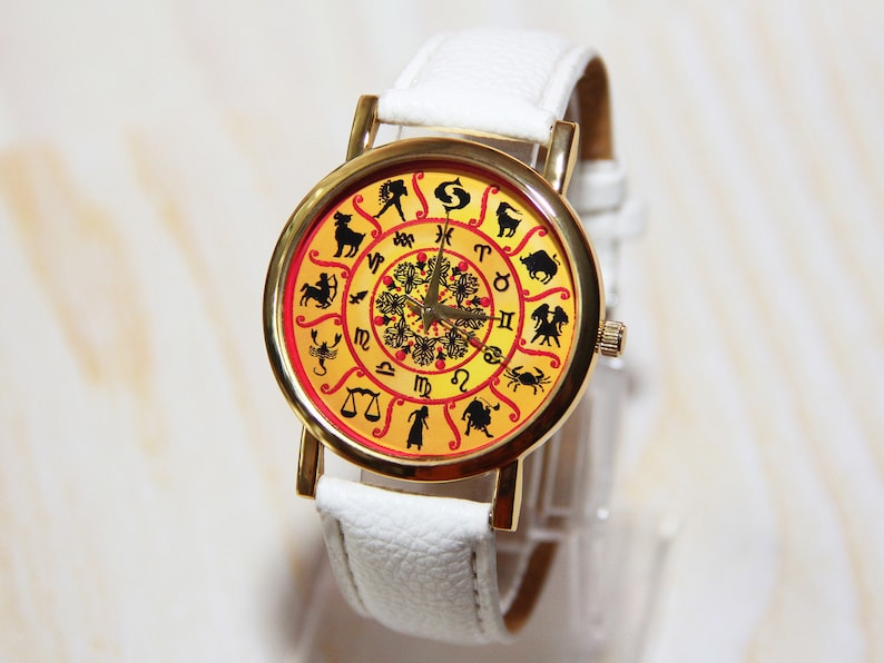 May include: A white wristwatch with a gold-tone case and a yellow face. The face features a zodiac design with black silhouettes of the astrological signs.