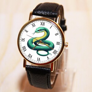 Puede incluir: Un reloj de pulsera de cuero negro con una esfera blanca y detalles dorados. La esfera presenta una serpiente verde con detalles amarillos y marcadores de hora en n&uacute;meros romanos.