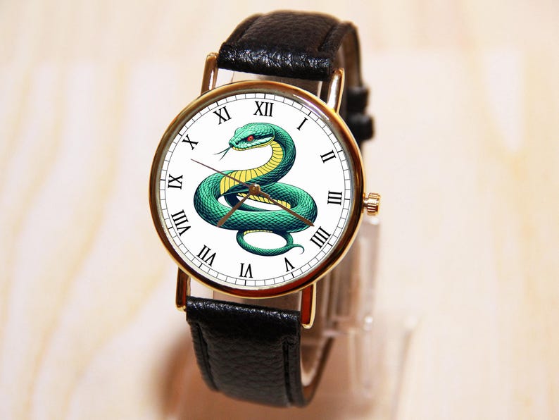 Puede incluir: Un reloj de pulsera de cuero negro con una esfera blanca y detalles dorados. La esfera presenta una serpiente verde con detalles amarillos y marcadores de hora en n&uacute;meros romanos.