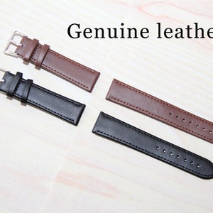 Puede incluir: Dos correas de reloj, una marr&oacute;n y otra negra, hechas de cuero genuino. Ambas correas tienen costuras y se muestran sobre una superficie de madera clara.
