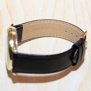Op de afbeelding: Een goudkleurig horloge met een zwarte leren band. De band is gemarkeerd met "20 GENUINE LEATHER".
