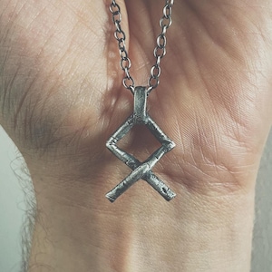 Puede incluir: Un collar de cadena de plata con un colgante de plata en forma de símbolo Vegvisir. El colgante está detallado con una superficie texturizada.