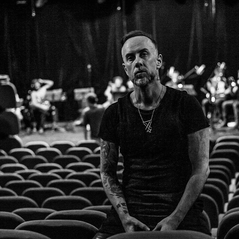 May include: A man with a beard and tattoos is sitting in a row of empty seats. He is wearing a black t-shirt with a silver necklace.