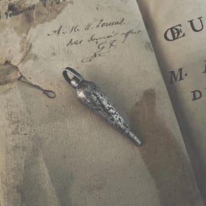 May include: A silver, textured, pointed pendant with a loop at the top. The pendant is lying on a piece of old, worn paper.