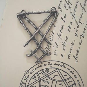 May include: A silver metal pendant in the shape of a triangle with a woven design. The pendant is on a white surface with handwritten text in French.