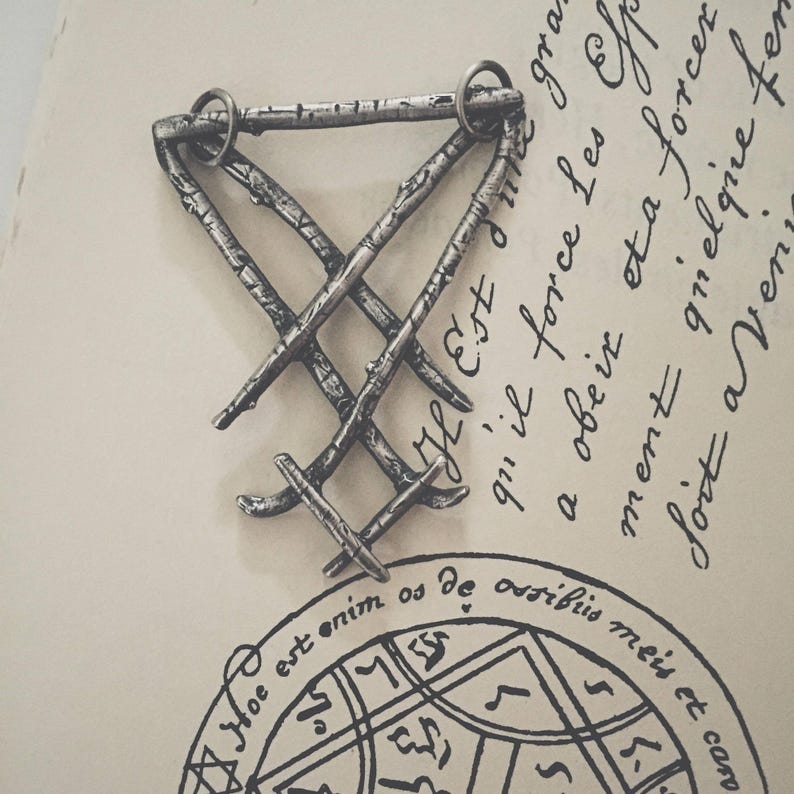 May include: A silver metal pendant in the shape of a triangle with a woven design. The pendant is on a white surface with handwritten text in French.