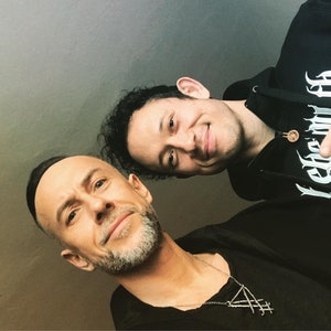 May include: Two men, one with a beard and a silver necklace, the other wearing a black hoodie with a graphic print, are smiling at the camera.