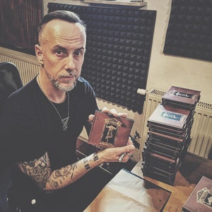 May include: A man with short dark hair and a beard is holding a book with a brown cover and a picture of a person on the front. The book is titled "Prophecies". There are several stacks of books behind him.