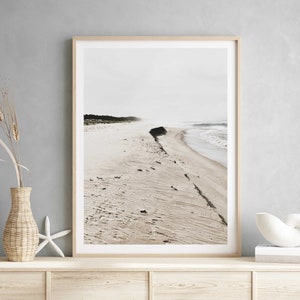 May include: A framed photograph of a beach scene with a long, narrow strip of sand leading to the ocean. The sand is light brown and the water is a light blue. The sky is a light gray. The photo is in a simple wooden frame.