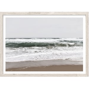 May include: A framed print of a seascape. The image features a beach with tan sand, foamy white waves, and dark teal water. The sky is a pale gray. The frame is a light, weathered wood.