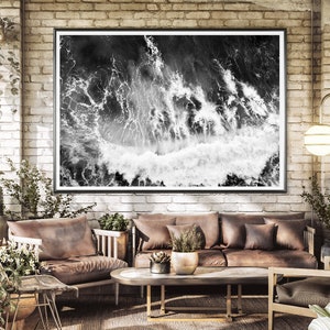 Puede incluir: Una fotograf&iacute;a en blanco y negro de olas rompiendo enmarcada en un marco negro cuelga de una pared de ladrillo en un ambiente de sala de estar. La habitaci&oacute;n cuenta con un sof&aacute; de cuero marr&oacute;n, una mesa de caf&eacute; y plantas.