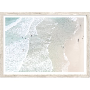 May include: An aerial view of a beach scene, framed in a light wood-tone frame. The image captures the ocean waves, people in the water, and the sandy shore. The water is a mix of turquoise and white, with the sand appearing light beige.
