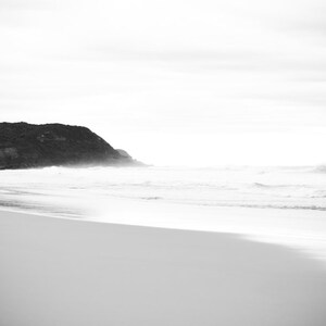 Op de afbeelding: Een zwart-witfoto van een strand met een rotsachtige klif op de achtergrond. Het zand is wit en het water is kalm.