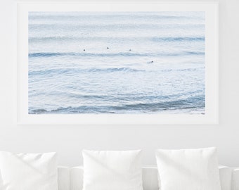 May include: A framed print of the ocean with surfers in the distance. The water is a light blue color with gentle waves. The print is in a white frame and is hanging above a white couch with white pillows.