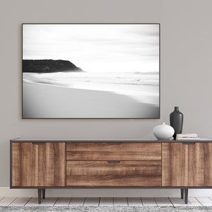 Op de afbeelding: Een zwart-witfoto van een strand met een rotsachtige klif op de achtergrond. De foto is ingelijst en hangt boven een houten consoletafel met twee laden en twee deuren. De consoletafel heeft een donkerbruine afwerking en zwarte metalen poten.