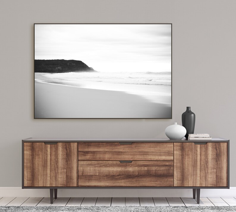 Op de afbeelding: Een zwart-witfoto van een strand met een rotsachtige klif op de achtergrond. De foto is ingelijst en hangt boven een houten consoletafel met twee laden en twee deuren. De consoletafel heeft een donkerbruine afwerking en zwarte metalen poten.