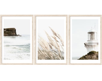 May include: A set of three framed prints featuring coastal scenes. The first print shows a rocky cliff and waves crashing on the shore. The second print features a close-up of tall, feathery grasses blowing in the wind. The third print shows a white lighthouse standing on a cliff overlooking the ocean.