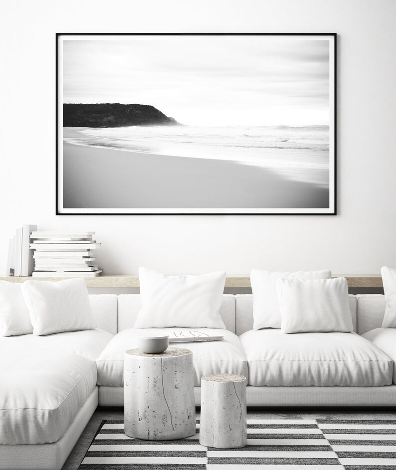 Op de afbeelding: Zwart-witfoto van een strand met een rotsachtige klif in de verte. De foto is ingelijst in een zwart frame en hangt aan een witte muur boven een witte bank.