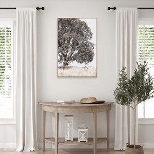 May include: A framed print of a black and white photograph of a tree with a light brown background. The print is hanging on a white wall above a wooden console table with a hat and two lanterns on top.