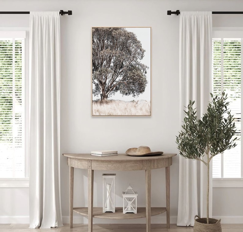 May include: A framed print of a black and white photograph of a tree with a light brown background. The print is hanging on a white wall above a wooden console table with a hat and two lanterns on top.