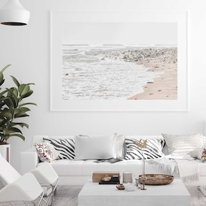 May include: A framed photograph of a beach scene with white sand, foamy waves, and gray rocks. The photo is hanging on a white wall above a white couch with black and white zebra print pillows and a white throw blanket.