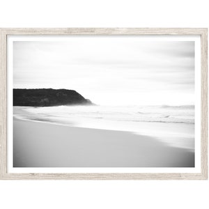 Op de afbeelding: Een zwart-witfoto van een strandsc&egrave;ne met een rotsachtige klif in de verte. De golven breken op de kust en het zand is glad en wit.