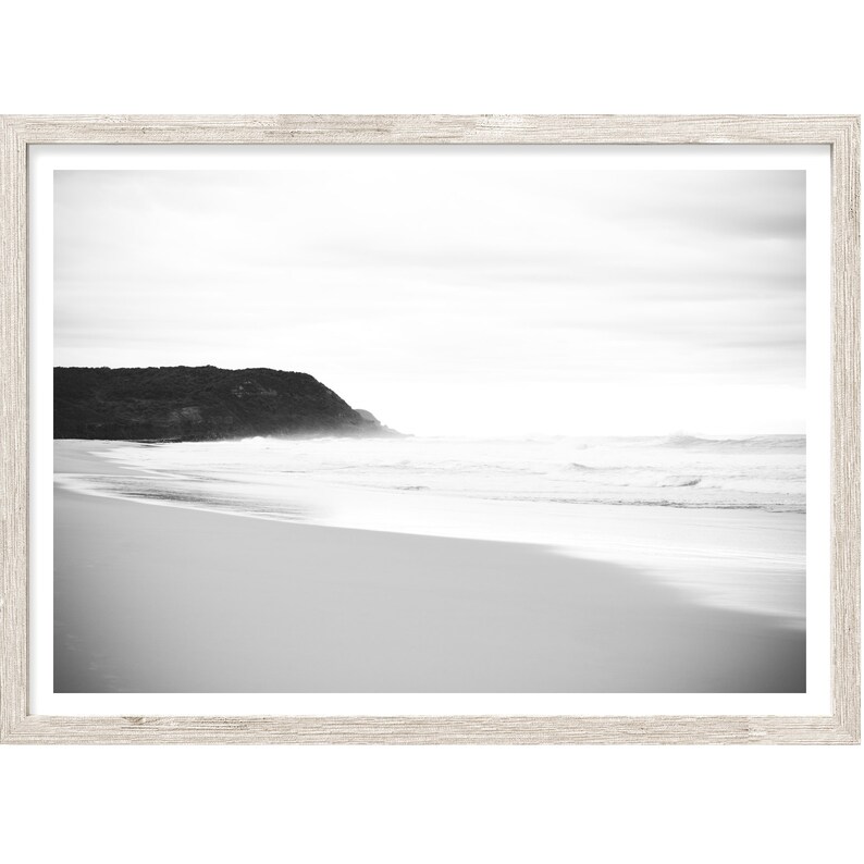 Op de afbeelding: Een zwart-witfoto van een strandsc&egrave;ne met een rotsachtige klif in de verte. De golven breken op de kust en het zand is glad en wit.