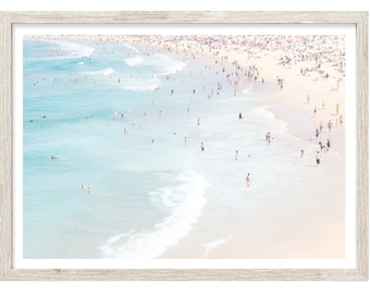 May include: A framed aerial photograph of a crowded beach with light blue ocean waves washing onto a pale sandy shore. Numerous small figures are scattered across the sand and in the water, creating a high-key, bright, and airy composition.