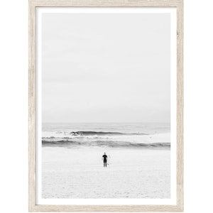 May include: Black and white photograph of a surfer standing on a beach with a surfboard under his arm, looking out at the ocean waves.