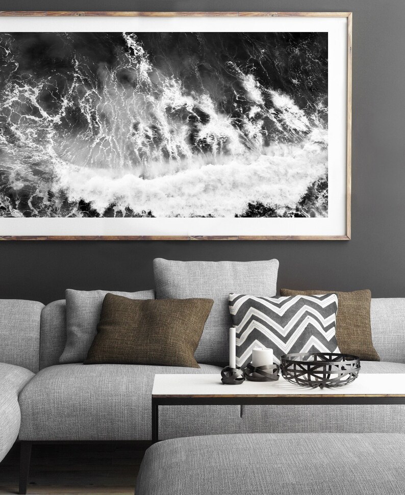 Puede incluir: Una fotograf&iacute;a en blanco y negro de olas del oc&eacute;ano rompiendo unas contra otras. La foto est&aacute; enmarcada y colgada en una pared gris oscuro sobre un sof&aacute; gris con almohadas de patrones marrones y blancos.