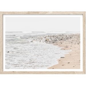 May include: A framed photograph of a beach scene. The photo shows a sandy beach with white foamy waves crashing on the shore. There are rocks and pebbles along the shoreline. The sky is a light blue and the water is a light blue-green color.