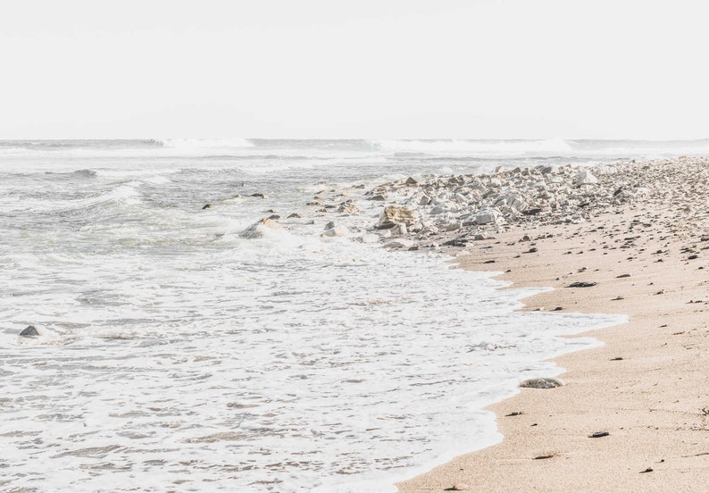 May include: A white sandy beach with foamy waves crashing on the shore. The water is a light blue and the sky is a pale white. The beach is lined with white rocks and the sand is a light brown.