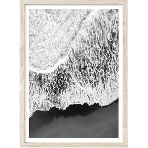 May include: Black and white aerial photograph of a wave crashing on a sandy beach. The wave is breaking into white foam and the sand is dark.