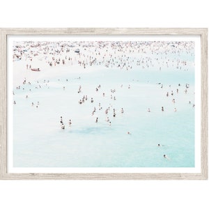 May include: A framed print featuring a beach scene with a multitude of people in the water and on the shore. The water is a light turquoise color, and the frame is a light wood color. A small boat is visible in the distance.