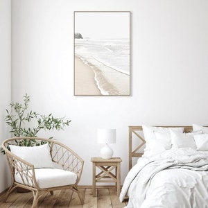 May include: A framed photograph of a beach scene with white sand and waves crashing on the shore. The photo is hanging on a white wall above a white bed with a white blanket and pillows.