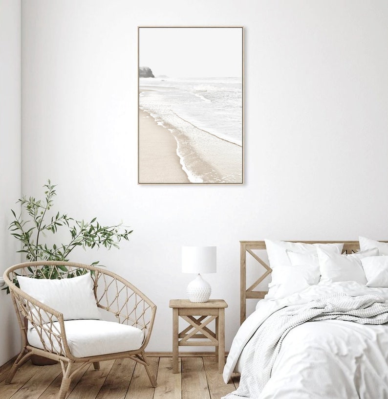 May include: A framed photograph of a beach scene with white sand and waves crashing on the shore. The photo is hanging on a white wall above a white bed with a white blanket and pillows.