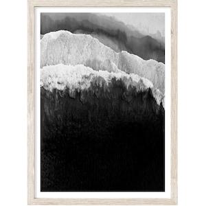 May include: A black and white photograph of ocean waves crashing against the shore. The image is taken from a high angle, looking down at the waves. The water is white and foamy, and the sand is dark and rocky.