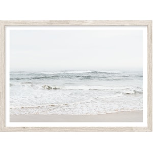 May include: A framed print featuring a serene coastal scene. The image captures the ocean with gentle waves breaking on a sandy beach. The sky is overcast, creating a soft, diffused light. The frame is a light, neutral wood tone, complementing the muted colors of the seascape.