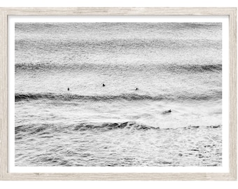 May include: Black and white photograph of a seascape with surfers in the water. The image shows a wide view of the ocean with rolling waves and a few surfers paddling on their boards.