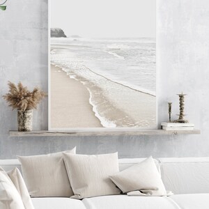 May include: A framed photograph of a beach scene with white sand, foamy waves, and a distant coastline. The photo is hanging on a white wall above a white couch with beige pillows.