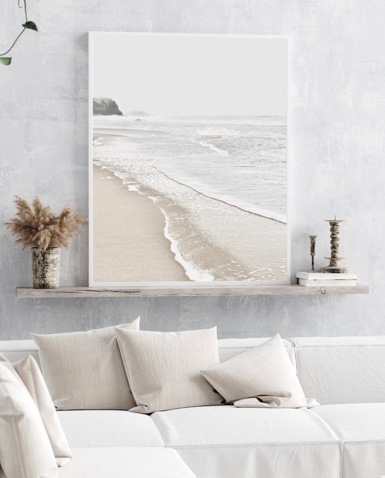 May include: A framed photograph of a beach scene with white sand, foamy waves, and a distant coastline. The photo is hanging on a white wall above a white couch with beige pillows.